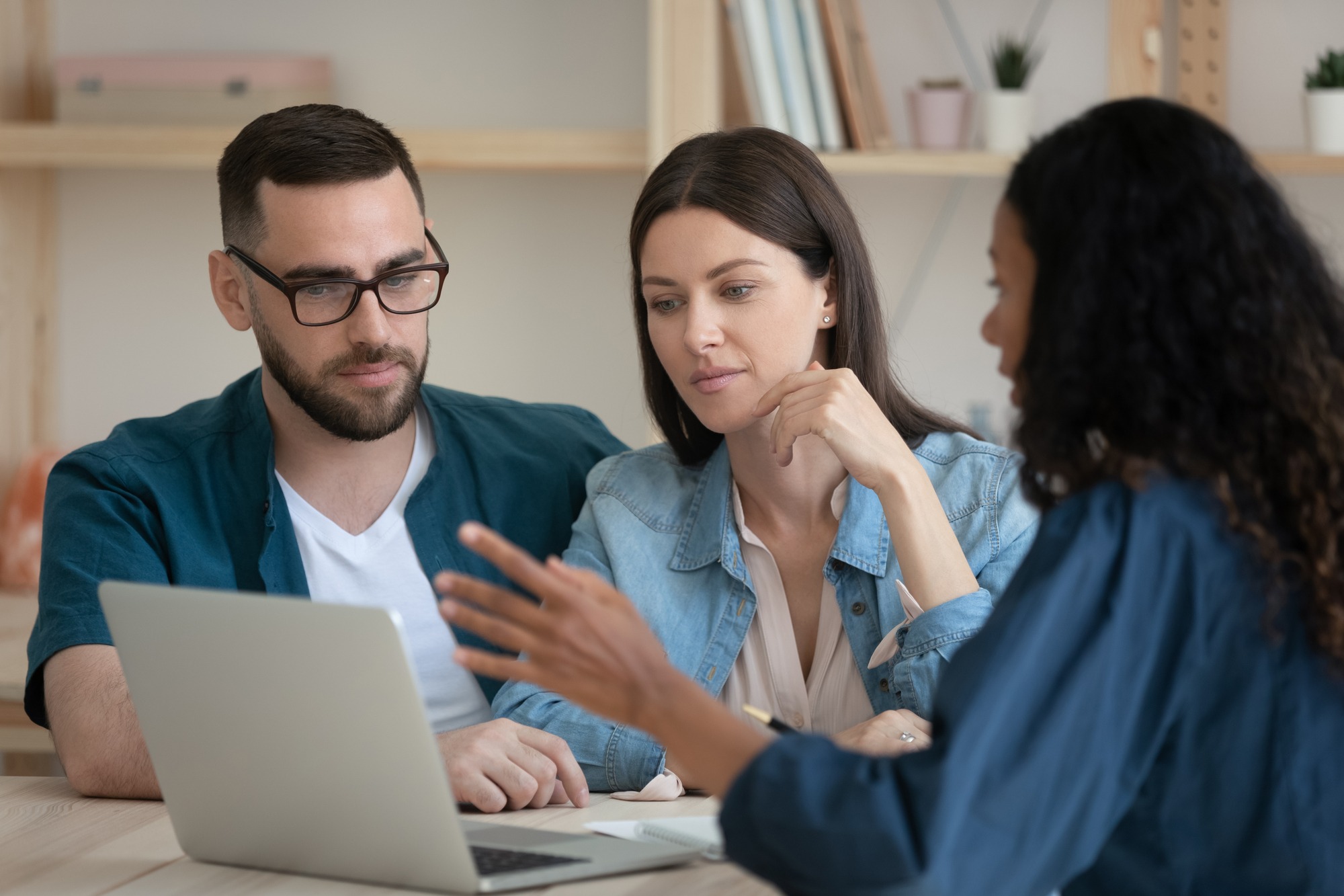 Focused young family couple listening to african american realtor at office. Mixed race financial advisor real estate agent lawyer showing service presentation on laptop to concentrated spouses.